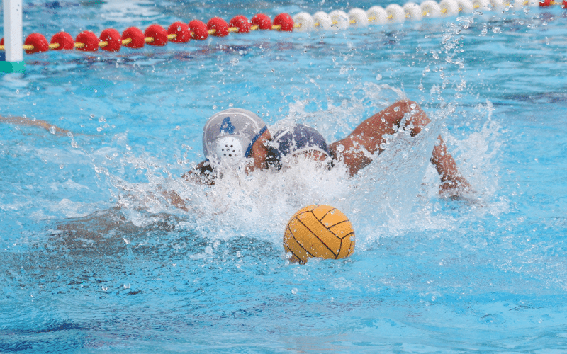 Players pass and shoot ball in water polo match, with referees and spectators on the sidelines